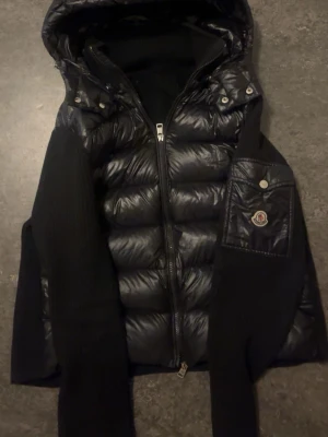 Svart dunjacka/cardigan från Moncler med huva - Snygg svart cardigan/dunjacka  från Moncler med glansig pufferlook, stickade ärmar och huva. Loggan scannar till moncler hemsida. Jackan har dragkedja framtill, quiltade partier och en Moncler-logga på ärmen. Perfekt för kalla dagar när du vill vara både varm och stilren.Priset kan sänkas vid snabb leverans,så tveka inte att skriva!