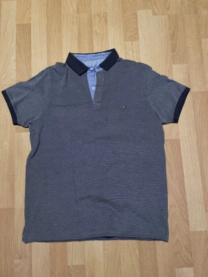 Grå pikétröja från Tommy Hilfiger - Grå pikétröja med mörkblå krage och ärmslut från Tommy Hilfiger. Klassisk passform med knappslå framtill och liten logga på bröstet. Tillverkad i mjukt bomullsmaterial som känns bekvämt och stilrent.