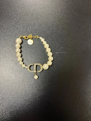 Dior armband med pärlor och guld - Lyxigt armband från Dior med vita pärlor och guldfärgade detaljer. Armbandet har en stor CD-logga i guld med små gnistrande stenar och en hängande pärla. Lås och bricka i guld med Dior-gravyr. Perfekt för dig som gillar exklusiva accessoarer.