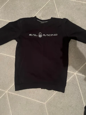 Svart Sail Racing sweatshirt - Svart sweatshirt från Sail Racing med vit logga och text på bröstet. Klassisk rund halsringning och ribbade muddar vid ärmslut och nederkant. Perfekt för en clean och sportig look.