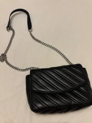 Svart quiltad axelväska med kedja - Snygg svart axelväska i quiltat konstläder med diagonal sömnad. Väskan har en silverfärgad kedjerem med en bekväm axelbit i svart läderimitation. Perfekt för dig som gillar en stilren och trendig look.