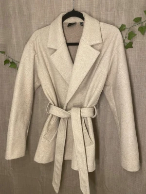 Beige kappa med knytskärp - Stilren beige kappa med bred krage och knytskärp i midjan. Kappan har två fickor framtill och är tillverkad i ett mjukt, ull-liknande material. Perfekt för kyligare dagar och ger en klassisk look.