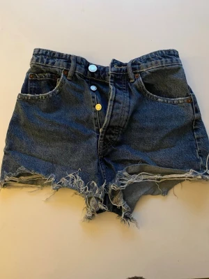 Mörkblå denimshorts med fransar - Säljer ett par mörkblå denimshorts med hög midja och råa fransade kanter. Shortsen har klassisk femficksdesign, knappar framtill och är tillverkade i bomull. Perfekta för en avslappnad och trendig sommarlook.