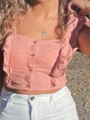 Rosa croppad blus med spetsdetaljer - Söt ljusrosa croppad blus med knappar framtill och breda axelband dekorerade med spets och volang. Toppen har fyrkantig ringning och är perfekt för varma dagar. Tillverkad i lätt bomullsmaterial som känns luftigt och bekvämt. Från H&M