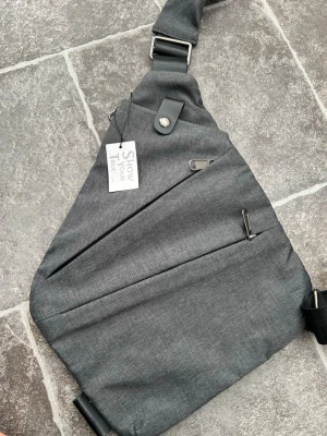 Grå crossbody väska med dragkedjor - Snygg grå crossbody ryggsäck med flera dragkedjefack och justerbar svart rem. Väskan har modern, trekantig form och detaljer i metall. Perfekt för dig som vill ha en stilren och praktisk väska till skolan eller stan.