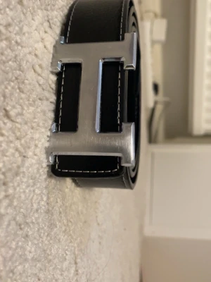 Svart skärp med silver H-spänne - Snyggt svart skärp från H med ett stort H-format spänne i silverfärgad metall. Skärpet är gjort i skinn och har vita kontrastsömmar längs kanterna för en clean och exklusiv look. Perfekt accessoar för att lyfta vilken outfit som helst.