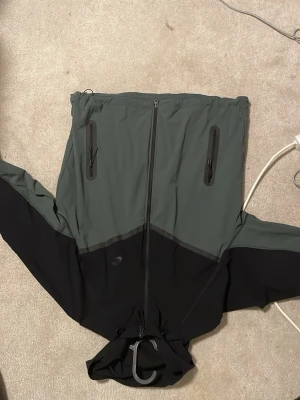 Grön och svart vindjacka med huva - Snygg vindjacka från Nike i tvåfärgad design, mörkgrön upptill och svart nedtill. Jackan har hel dragkedja framtill, två vertikala bröstfickor med dragkedja och justerbar huva. Tillverkad i lätt och slitstarkt material, perfekt för utomhusaktiviteter.