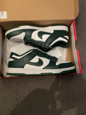 Nike Dunk Low  - Snygga Nike Dunk Low sneakers i grönt och vitt läder med klassisk Swoosh på sidan. Yttersulan är grön och ovandelen har perforerad tåbox för extra stil. Perfekta för dig som vill ha en clean och sportig look.