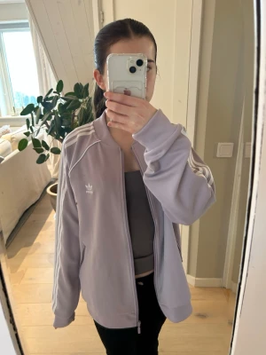 Ljuslila Adidas track jacket - Snygg ljuslila Adidas track jacket med dragkedja och klassiska vita ränder längs ärmarna. Jackan har två fickor med dragkedja och Adidas logga på bröstet. Perfekt för träning eller chill vardagsstil. Är i storlek 46 men skulle säga att den passar någon i S om man vill ha lite mer oversized look.