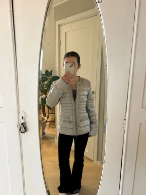 Ljusgrå pufferjacka med volangdetalj - Snygg ljusgrå pufferjacka med raka linjer och volangdetalj längs dragkedjan. Jackan har två fickor med dragkedja och är lätt vadderad för extra värme. Perfekt för kalla dagar och enkel att matcha med olika outfits. Den är i storlek L men skulle säga att den mer är S/M.