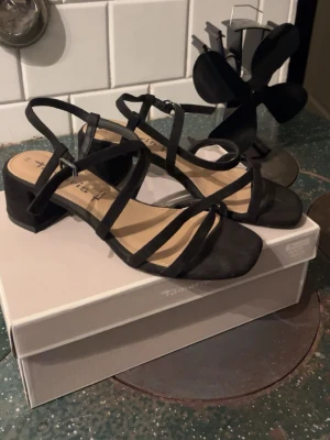Svarta sandalettpumps från Tamaris - Snygga svarta skor från Tamaris med låg, bred klack och tunna remmar över foten. Skorna har öppen tå och justerbart spänne. Har endast använt en gång på balen. Super sköna att gå i pga den bredare klacken.