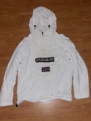 Vit jacka från Napapijri - Säljer en vit vindjacka från Napapijri med huva och dragkedja vid halsen. Jackan har en stor magficka med Napapijri-logga samt patch på ärmen. Perfekt för blåsiga dagar och i skidbacken.