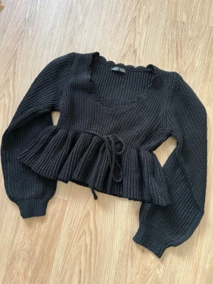 Svart stickad topp med knytning och v-ringning, strl S - Svart snygg stickad topp med knytband i midjan och bred volang nedtill. Djup v-ringning och lösare ärmar. Sparsamt använd, bra skick!