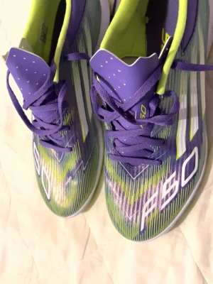 Adidas F50 lila fotbollsskor - Säljer ett par Adidas F50 fotbollsskor i lila med vita och neongröna detaljer. Skorna har snörning, låg profil och klassisk design med tre vita ränder på sidan. Insidan är fodrad i neongrönt och ovandelen är i syntetmaterial för bra bollkontroll.