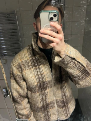 Beige och brun rutig cardiganjacka från Abercrombie & Fitch - Snygg cardiganjacka i beige och brunt rutigt mönster från Abercrombie & Fitch med dragkedja framtill och hög krage. Jackan har en lite boxig passform och är tillverkad i ett ull-liknande material som ger en mysig och trendig look.