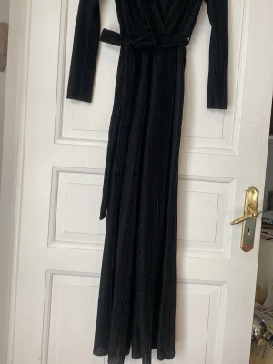 Svart glittrig jumpsuit från NA-KD - Snygg svart jumpsuit från NA-KD med glittereffekt, djup omlott-ringning och långa ärmar. Plagget har ett knytband i midjan och vida ben. Materialet är ribbat och stretchigt, vilket ger en bekväm och trendig look.