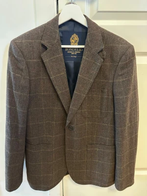 Brun rutig kavaj från Bondelid - Stilren brun kavaj från Bondelid med rutmönster i beige och rött. Klassisk modell med två framfickor, bröstficka och två knappar. Fodrad insida och snyggt skuren krage.