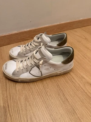 Vita Philippe Model sneakers med mockadetaljer - Snygga vita sneakers från Philippe Model med gråa mockadetaljer och olivgröna partier vid hälen. Klassisk låg modell med vita snören och diskret logga på sidan. Perfekta för en clean och avslappnad stil.