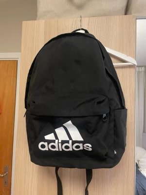 Svart ryggsäck från Adidas - Säljer en klassisk svart ryggsäck från Adidas med stort vitt logotryck på framsidan. Ryggsäcken har ett stort huvudfack, en framficka med dragkedja och två sidofickor. Tillverkad i slitstarkt tyg och har justerbara axelband.