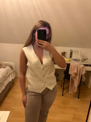 Krämvit väst med knappar - Stilren off white/kräm vit väst med djup v-ringning och knäppning framtill. Västen har en klassisk passform och är perfekt att styla över en skjorta eller t-shirt. Snygg och enkel design som passar till många olika outfits. En present som inte riktigt är något för mig. Den är något stor i storlek den är i ”regular fit”. Helt oanvänd med prislapp kvar.