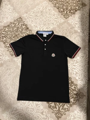 Svart pikétröja från Moncler - Klassisk svart pikétröja från Moncler med vit och röd rand på krage och ärmslut. Tröjan har knappslå med tre vita knappar och Moncler-logga broderad på bröstet. Perfekt för en stilren och sportig look.