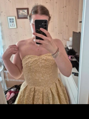 Gul axelbandslös kortklänning - Söt axelbandslös kortklänning i gul nyans med blommigt mönster. Klänningen har en utställd kjol och markerad midja, perfekt för varma dagar. Materialet känns som bomull och ger en luftig känsla.