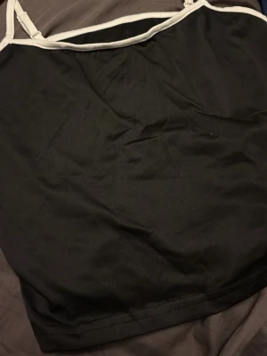 Svart linnetopp med vita band - Snygg svart linnetopp med kontrasterande vita band längs kanterna och tunna axelband. Perfekt att styla med jeans eller kjol för en clean och enkel look. Materialet känns mjukt och lätt mot huden.