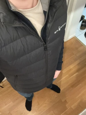 Svart dunväst från Sail racing - Snygg svart dunväst från Sail Racing med quiltad design och dragkedja framtill. Västen har hög krage och diskret logga på bröstet. Perfekt att slänga över en hoodie eller tröja för extra värme. Klassisk och stilren look som funkar till det mesta. Pris kan diskuteras och skriv gärna frågor💴