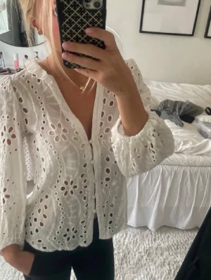 Vit broderad blus  - Säljer en vit blus med broderade hålmönster och puffiga långärmar. Blusen har v-ringning och knäppning framtill. Materialet är lätt och luftig bomull, perfekt för en trendig och avslappnad look.