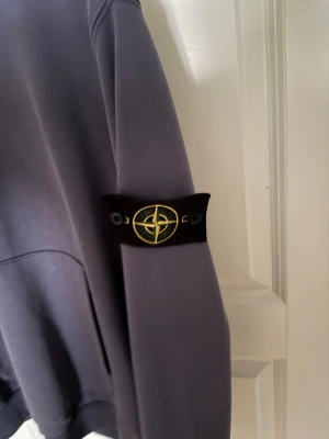 Grå hoodie från Stone Island - Säljer en stilren grå hoodie från Stone Island med klassisk patch på vänster ärm. Hoodien har magficka framtill, ribbade muddar och är tillverkad i mjuk bomull. Perfekt för dig som gillar streetwear och vill ha något bekvämt men ändå snyggt.