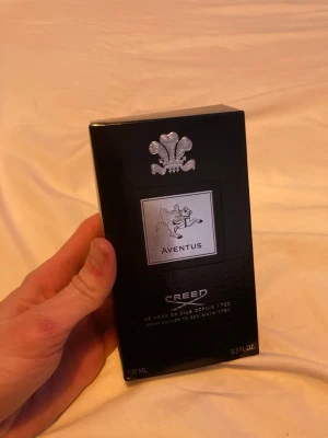 Creed Aventus Eau de Parfum 100 ml - Lyxig parfym från Creed, Aventus, i en elegant svart kartong med silverdetaljer och logotyp. Förpackningen rymmer 100 ml och har en exklusiv känsla med tydlig märkning på både fram- och baksida. Perfekt för dig som gillar klassiska och sofistikerade dofter.