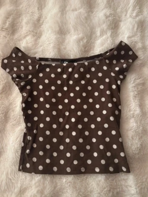 Brun prickig offshoulder topp - Snygg brun offshoulder topp med vita prickar. Toppen har en figurnära passform och är perfekt för dig som gillar retrovibbar. Tillverkad i mjukt bomullsmaterial som känns skönt mot huden. Passar till både jeans och kjol för en trendig look.