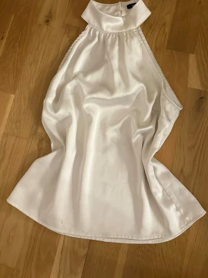 Vit halterneck topp i satin - Snygg vit halterneck topp i glansigt satinmaterial med hög krage och ärmlös design.  knäpps i nacken. Dock har den en liten brun fläck längst ner! Dock inte man tänker på