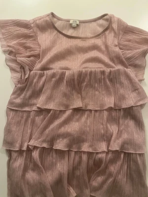 Glittrig rosa volangklänning River Island - Supersöt rosa klänning från River Island med glittrigt tyg och flera volanglager. Klänningen har korta, luftiga ärmar och rund halsringning. Perfekt för dig som vill sticka ut med en feminin och trendig look.