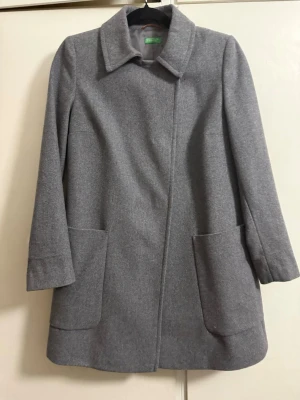 Grå klassisk rock från Benetton - Stilren grå rock från Benetton med rak passform och stora fickor framtill. Rocken har klassisk krage och är tillverkad i ett mjukt, ull-liknande material. Perfekt för dig som gillar enkel och tidlös design.