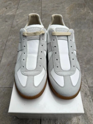  Maison margiela gats.  - Snygga sneakers i vitt och ljusgrått med klassisk design och gummisula. Ovandelen är i en mix av läder och mocka, vilket ger en stilren och fräsch look. Perfekta för dig som gillar minimalistisk stil och vill ha något som funkar till det mesta.