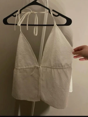 Vit broderad linnetopp med knyt - Säljer en söt vit linnetopp med broderade detaljer och knyt i nacken samt under bysten. Toppen har v-ringad hals och är ärmlös, perfekt för varma dagar. Lätt och luftig med en feminin touch.