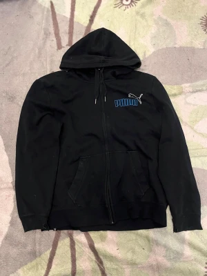 Blå hoodie med dragkedja från Puma - Blå hoodie från Puma med dragkedja framtill och blått Puma-tryck på bröstet. Klassisk huva med dragkedja och känguruficka. Perfekt för en avslappnad streetstyle-look. Tillverkad i mjukt bomullsmaterial.