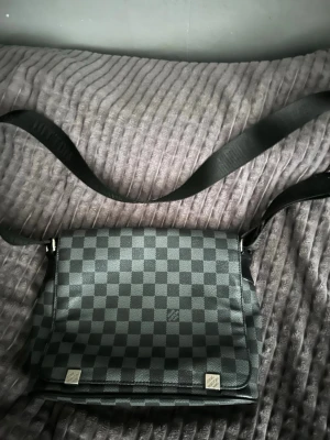 Louis Vuitton axelväska Damier Graphite - Snygg axelväska från Louis Vuitton i klassiskt Damier Graphite-mönster. Väskan är svart och grå med justerbar axelrem och lock. Tillverkad i canvas med detaljer i läder och metall. Perfekt för dig som vill ha en stilren och ikonisk väska.
