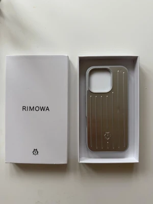 RIMOWA silver mobilskal i aluminium, Iphone 16 Pro - Snyggt och stilrent mobilskal från RIMOWA i silverfärgat aluminium med klassiska räfflade linjer. Skalets insida är grå och har märkets logga tryckt. Passar perfekt för dig som vill ha ett exklusivt och tåligt skydd till din mobil.