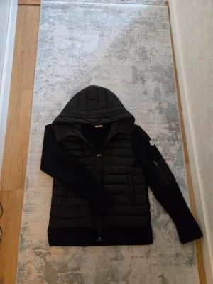 Svart pufferjacka med huva Moncler - Snygg svart pufferjacka från Moncler med vadderad front och huva. Jackan har stickade ärmar och en Moncler-logga på ena ärmen samt dragkedja framtill. Perfekt för kalla vinterdagar och riktigt trendig look. Priset är inte fastpris och kan sänkas i snabbaffär om det äe seriösa köpare. Kan försöka hitta kvitton om önskas!