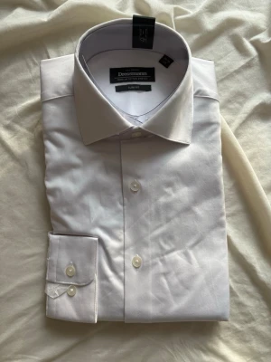 Vit slim fit skjorta från Dressmann - Klassisk vit skjorta från Dressmann i slim fit-modell. Tillverkad i premium bomull med stretch för extra komfort. Skjortan har långa ärmar, knappar framtill och en stilren, tidlös design som passar perfekt till skjorta och slips.