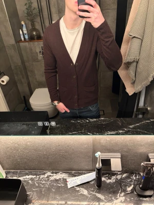 Brun kofta från LOGG - Säljer en stilren brun kofta med knäppning framtill och två fickor. Koftan har en smal passform och är perfekt att slänga över en t-shirt. Materialet känns mjukt och stretchigt, vilket gör den skön att bära hela dagen.