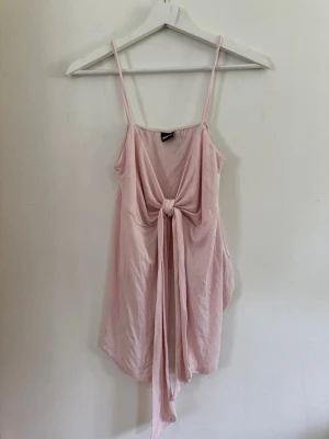 Ljusrosa kort klänning med knyt M - Söt ljusrosa kort klänning med tunna axelband och knyt framtill. Klänningen har en avslappnad passform och är perfekt för varma dagar. Tillverkad i mjukt tyg som känns skönt mot huden.