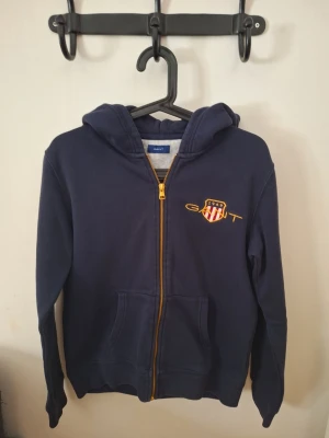 Marinblå hoodie från GANT med dragkedja - Mörkblå hoodie från GANT med gul dragkedja och broderad logga på bröstet. Klassisk huva, känguruficka framtill och mjukt bomullsmaterial. Perfekt för chill dagar och snygg streetstyle. Passar ungefär 160-170 cm 