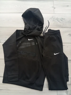 Svart Nike set med hoodie och byxor - Säljer ett svart set från Nike med hoodie och matchande byxor. Hoodien har dragkedjeficka och tryckt Nike-logga på bröstet, byxorna har resår i midjan och vit Nike-logga. Snygg och sportig look med bekväma material och detaljer.