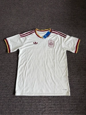 Fotbollströja  - Säljer en vit Spanien fotbollströja från Adidas med vinröda detaljer och gula inslag vid kragen. Tröjan har korta ärmar, Adidas klassiska tre ränder på axlarna och Spaniens landslagsemblem på bröstet. Tillverkad i lätt och funktionellt material.