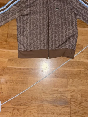 Brun ziptröja från Lacoste - Snygg brun ziptröja från Lacoste med diskret mönster och klassisk krokodillogga på bröstet. Tröjan har ribbade muddar, hel dragkedja och vita samt blå ränder längs ärmarna. Perfekt för en sportig och trendig look.