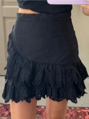 Svart volangkjol med broderade detaljer - Supersnygg svart kortkjol med volanger och broderade detaljer. Kjolen har en omlott-look och är perfekt för dig som gillar en feminin och trendig stil. Tillverkad i bomull för en skön känsla och snygg passform.