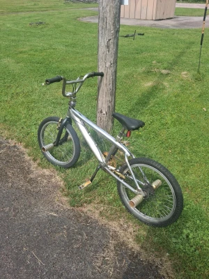 Silver BMX-cykel med pegs - Säljer en silverfärgad BMX-cykel med svart sadel och svarta handtag. Cykeln har pegs på både fram- och bakhjul, perfekt för tricks och street riding. Ramen är i metall och pedalerna är i plast med metallkant. Inga synliga märken på cykeln.